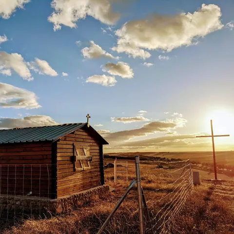 Caseta de madera con una cruz en el tejado y una gran cruz en el campo, al atardecer