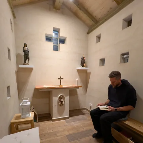 Interior sencillo de la capilla, con altar, crucifijo y dos imágenes; un hombre lee sentado en un banco
