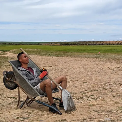 Un joven descansa recostado en una carretilla, con la pala en las manos, frente a los campos